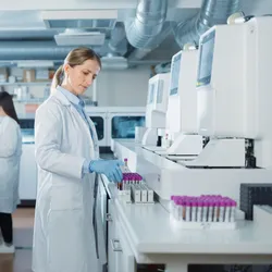 Biomedical Researchers Conduct Experiments in a Modern Laboratory. Female Scientist Preparing a Set of Samples in Test Tubes with Purple Caps. Colleague Analyzes Data on a Computer in the Background