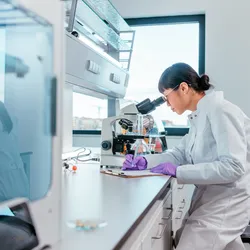 Gloved female scientist using a microscope in a lab, highlighting the importance of glove fit and dexterity in thin-mil glove selection.