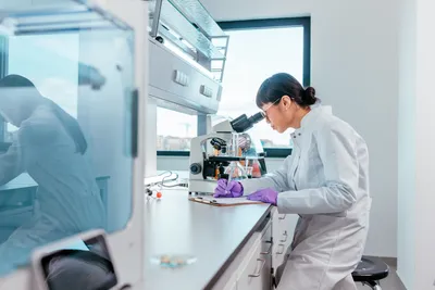 Gloved female scientist using a microscope in a lab, highlighting the importance of glove fit and dexterity in thin-mil glove selection.