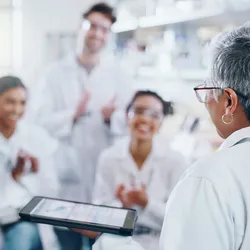 Senior scientist presenting data to a group of motivated lab staff applauding in a lab setting.