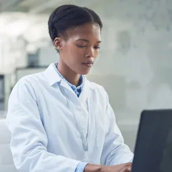 A black woman in a lab coat types on a laptop computer