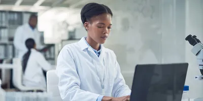 A black woman in a lab coat types on a laptop computer