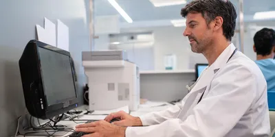 Male healthcare professional in a white lab coat working on a desktop computer in a clinical or laboratory office environment, focused on managing or analyzing data.