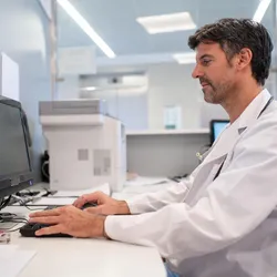Male healthcare professional in a white lab coat working on a desktop computer in a clinical or laboratory office environment, focused on managing or analyzing data.