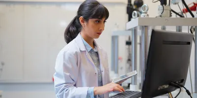 Female scientist in a lab coat working on a desktop computer while holding a tablet, focused on data entry or analysis in a laboratory environment with technical equipment in the background