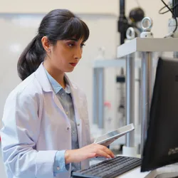 Female scientist in a lab coat working on a desktop computer while holding a tablet, focused on data entry or analysis in a laboratory environment with technical equipment in the background
