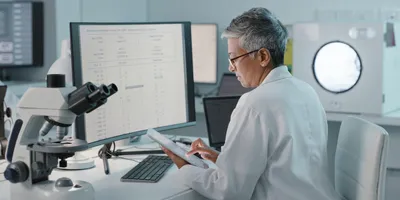 Scientist in a lab coat using a tablet while analyzing genetic or clinical data displayed on a large computer monitor in a laboratory setting, with a microscope in the foreground