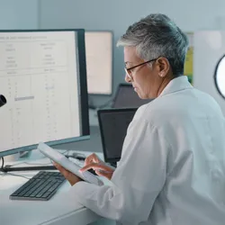 Scientist in a lab coat using a tablet while analyzing genetic or clinical data displayed on a large computer monitor in a laboratory setting, with a microscope in the foreground