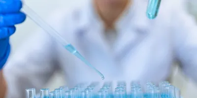 Gloved technician pipetting into vial rack during batch sample preparation