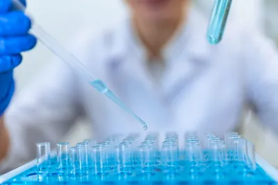 Gloved technician pipetting into vial rack during batch sample preparation