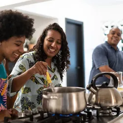 A joyful family scene in a kitchen, with a woman and girl smiling and cooking at a stove, and a man in the background. The atmosphere is warm and lively.