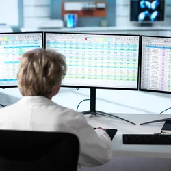 View from behind of a woman with short blonde hair in a lab coat, in front of a triple monitor desktop covered displaying spreadsheets