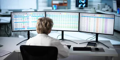 View from behind of a woman with short blonde hair in a lab coat, in front of a triple monitor desktop covered displaying spreadsheets