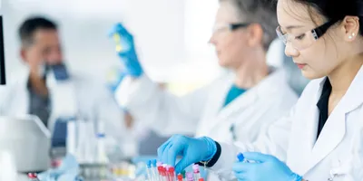 A medical lab technician is seen sorting plasma samples as a small team works together to analyze the samples. They are each wearing lab coats, protective eyewear and gloves as they work diligently.