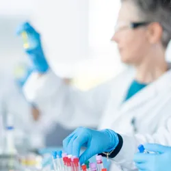 A medical lab technician is seen sorting plasma samples as a small team works together to analyze the samples. They are each wearing lab coats, protective eyewear and gloves as they work diligently.