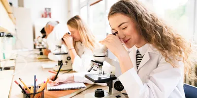 Students in an academic lab using microscopes and taking notes, illustrating resource constraints and hands-on learning in educational laboratories.