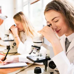 Students in an academic lab using microscopes and taking notes, illustrating resource constraints and hands-on learning in educational laboratories.