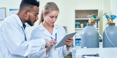Two lab professionals discussing information on a tablet standing next to lab gas canisters and equipment