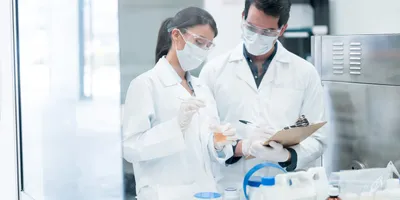 Two scientists in lab coats, face masks, and safety goggles working with a chemical sample in a laboratory, illustrating lab safety protocols and hazard awareness.