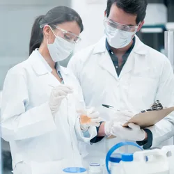 Two scientists in lab coats, face masks, and safety goggles working with a chemical sample in a laboratory, illustrating lab safety protocols and hazard awareness.