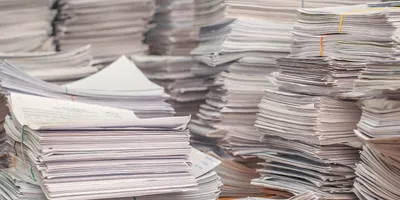 Stacks of paper cover a desk, representing slow and inefficient paper-based lab workflows