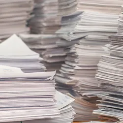 Stacks of paper cover a desk, representing slow and inefficient paper-based lab workflows