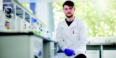 A young scientist in a lab coat and gloves sits at a lab bench facing the camera
