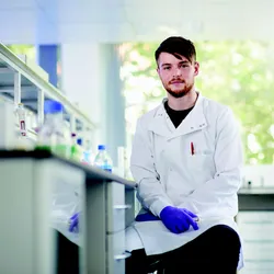 A young scientist in a lab coat and gloves sits at a lab bench facing the camera
