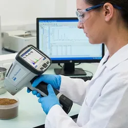 Scientist using a handheld XRF analyzer to test a prepared soil sample for heavy metals in an environmental laboratory, with spectral data displayed on a background monitor.