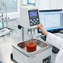 A female scientist in a lab coat and safety glasses operates a rotational viscometer, which is testing a red sauce sample submerged in a water bath, with a computer screen displaying data in the background.