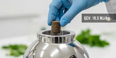 Photorealistic close-up of a stainless steel bomb calorimeter vessel being loaded by a gloved hand. The sample inside is a compressed pellet of biomass. Illustrative of the role of calomirmetry in crop and soil science