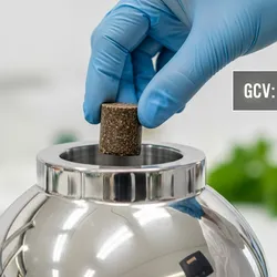 Photorealistic close-up of a stainless steel bomb calorimeter vessel being loaded by a gloved hand. The sample inside is a compressed pellet of biomass. Illustrative of the role of calomirmetry in crop and soil science