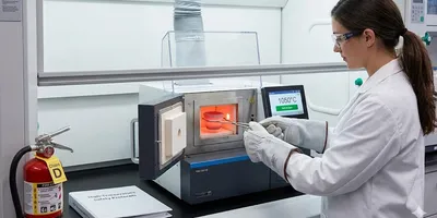 Scientist wearing heat-resistant PPE and safety glasses uses tongs to carefully place a crucible into a glowing thermal analysis instrument furnace, illustrating high-temperature lab safety protocols and engineering controls.