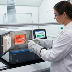 Scientist wearing heat-resistant PPE and safety glasses uses tongs to carefully place a crucible into a glowing thermal analysis instrument furnace, illustrating high-temperature lab safety protocols and engineering controls.