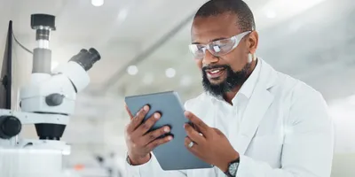 A scientist in a lab coat and safety glasses smiles while looking at a digital tablet