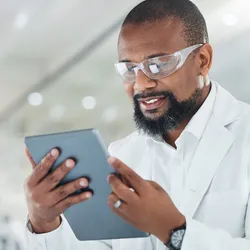A scientist in a lab coat and safety glasses smiles while looking at a digital tablet