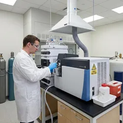 Scientist wearing PPE maintains a mass spectrometer in a safety-compliant lab featuring proper ventilation, secured gas cylinders, and high-voltage warning signage