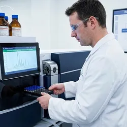 A scientist in a modern laboratory loads sample vials into a high-tech mass spectrometer while monitoring a chromatogram on the display screen, with a regulatory validation framework chart visible on the wall in the background.