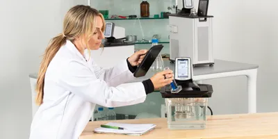 A lab technician in a white coat operates a PolyScience PolyTemp circulator, holding a tablet and a graduated cylinder with blue liquid in a modern laboratory