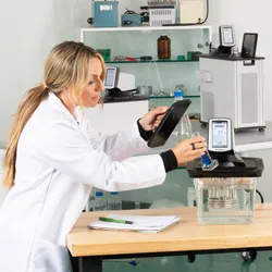 A lab technician in a white coat operates a PolyScience PolyTemp circulator, holding a tablet and a graduated cylinder with blue liquid in a modern laboratory