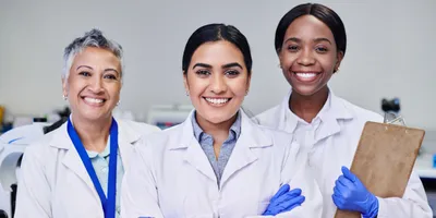 Three women in lab coats smiling together, representing women in leadership.