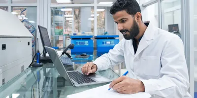 Young male biochemist using laptop while writing on clipboard at laboratory