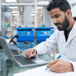 Young male biochemist using laptop while writing on clipboard at laboratory