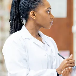 A focused Black woman in a white lab coat stands in a professional setting, writing on a clipboard while examining shelves of medications, signaling concentration, accuracy, and scientific work.