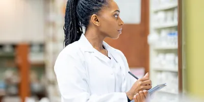 A focused Black woman in a white lab coat stands in a professional setting, writing on a clipboard while examining shelves of medications, signaling concentration, accuracy, and scientific work.
