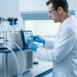 A laboratory professional in proper PPE operates a freeze-dryer system connected to a vacuum pump in a clean, modern food science facility.