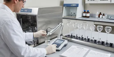 A laboratory QA/QC scene showing a scientist in a white lab coat using a bench-top conductivity meter to test a sample of rinse water drawn from a stainless steel automated glassware washer.