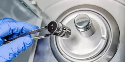 A operator's hand is holding steel calibration weight to place on the analytical balance.