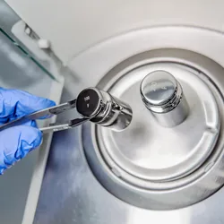 A operator's hand is holding steel calibration weight to place on the analytical balance.