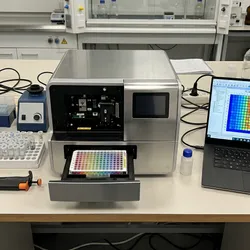 A top-down view of a modern multi-mode microplate reader on a laboratory bench with a 384-well plate loaded in the instrument drawer, flanked by a multichannel pipette, labeled sample tubes in a rack, and a laptop monitor displaying a color-coded fluorescence intensity heatmap.